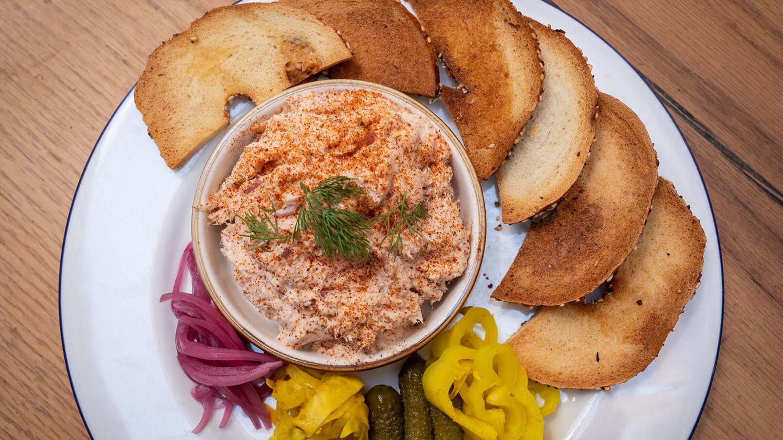A bowl of fish dip on a plate surrounded by slices of toasted bread and pickled vegetables